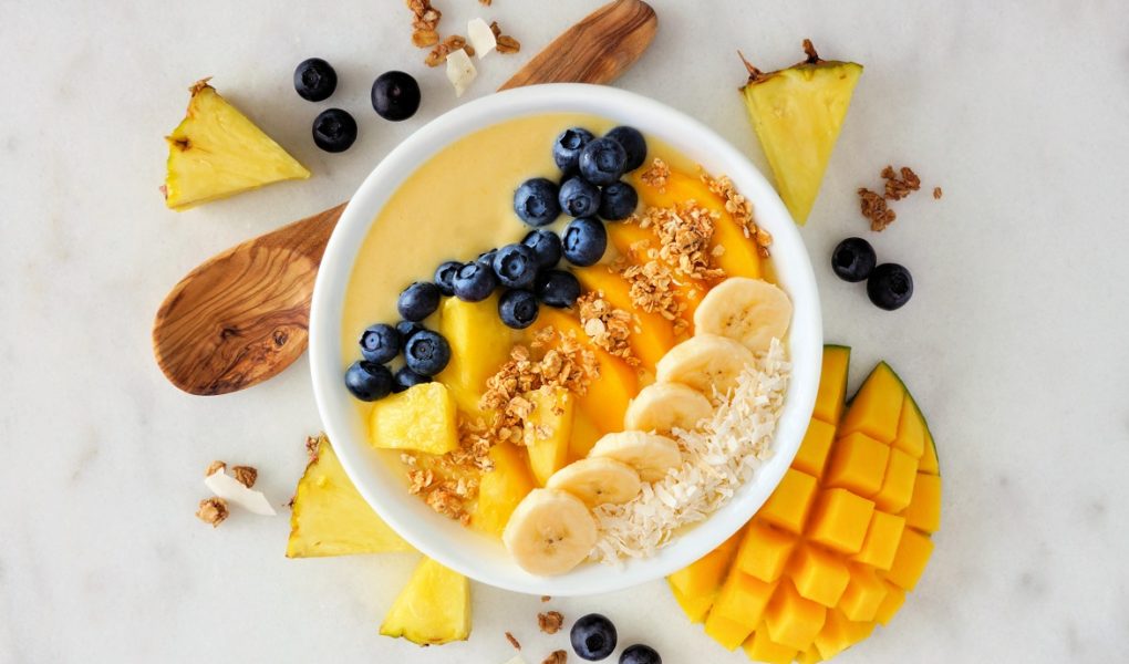 Healthy pineapple, mango smoothie bowl with coconut, bananas, blueberries and granola. Above view scene on a bright background. Czym są smoothie bowl? Pięknie skomponowany posiłek, serwowany w głębokich miseczkach